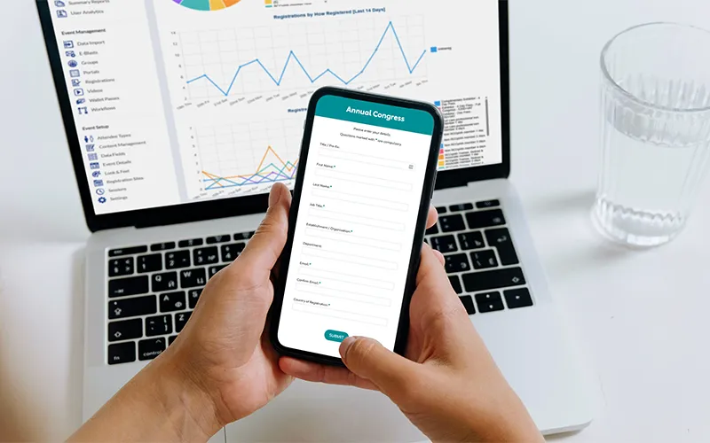 Person using a registration site on a smartphone, with registration software on a laptop screen in the background